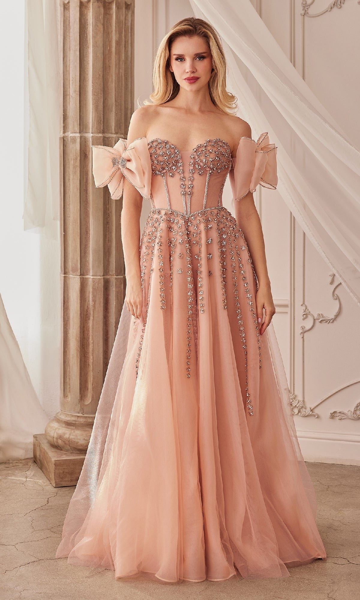 A woman stands in an elegant room wearing the Andrea and Leo A1338 strapless long prom dress, a blush pink gown adorned with sparkling beaded embellishments and tulle accents.